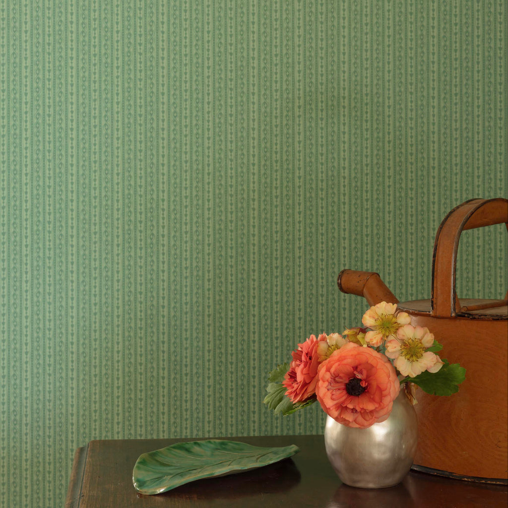 Green patterned wallpaper shown on a wall, with a small table in the foreground holding a vase of flowers and a brown watering can.