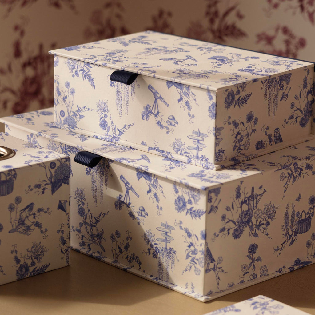 A set of decorative storage boxes with a blue and white toile de Jouy pattern, arranged on a beige wooden vanity table against a matching floral wallpaper background. Includes tissue box and a5 storage box.