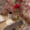 A styled tabletop scene with a blue-and-white toile patterned tray holding a metallic bird sculpture and a small card, alongside a matching patterned cup with red pens and a glass vase of pink flowers against floral wallpaper. A Delft Toile De Jouy pencil holder is the showcase piece.