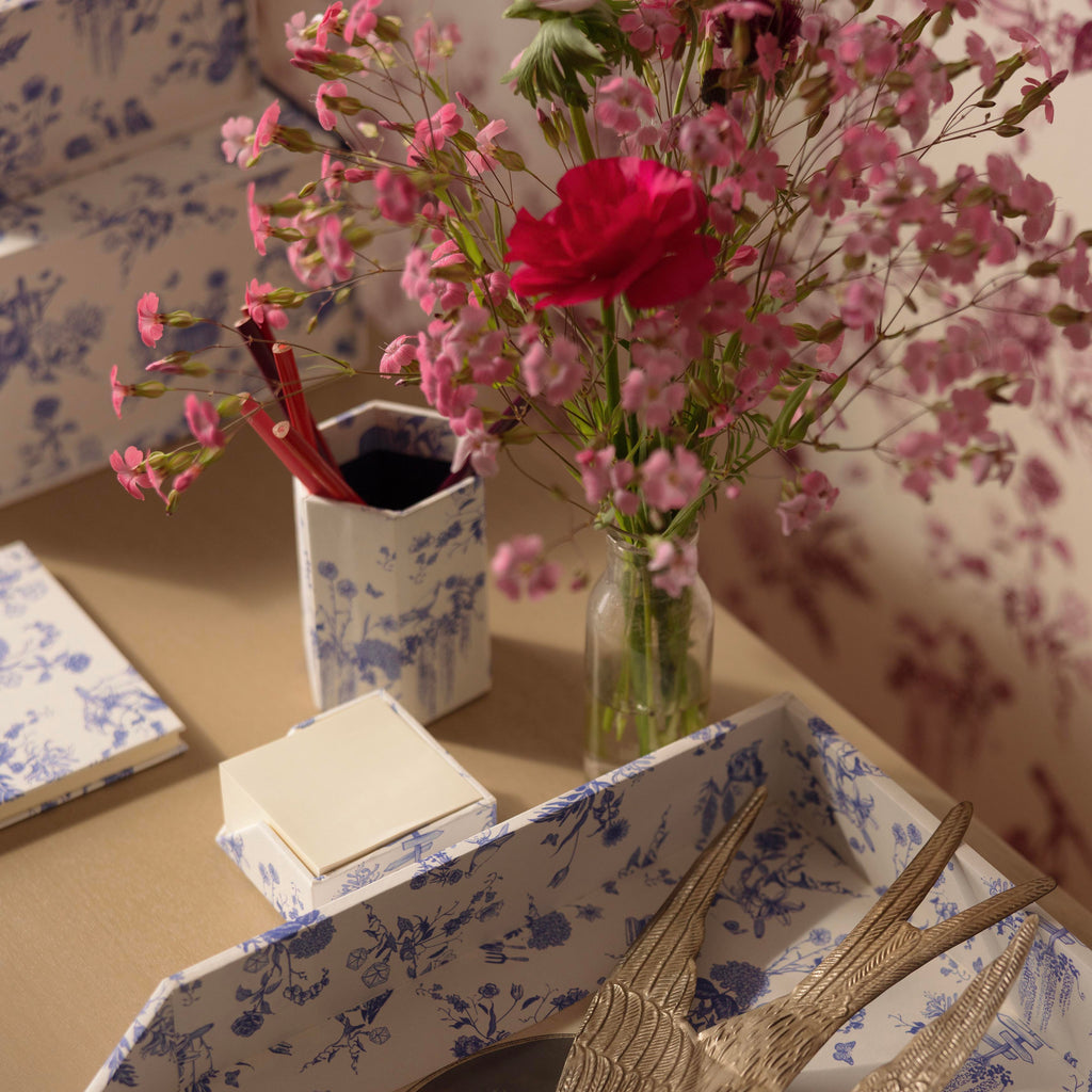 A styled tabletop scene with a blue-and-white toile patterned tray holding a metallic bird sculpture and a small card, alongside a matching patterned cup with red pens and a glass vase of pink flowers against floral wallpaper. A Delft Toile De Jouy pencil holder is the showcase piece.