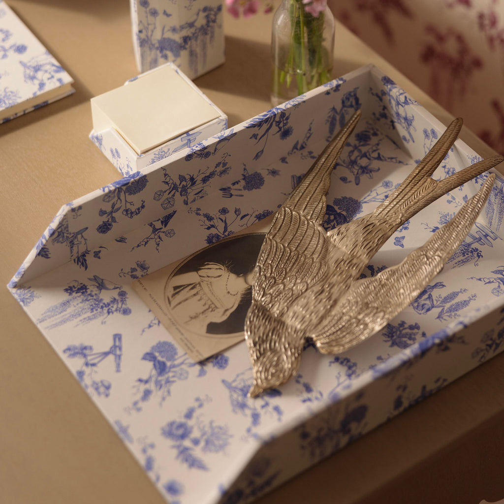 A styled tabletop scene with a blue-and-white Delft Toile De Jouy patterned letter paper tray holding a metallic bird sculpture and a small card, alongside a matching patterned cup with red pens and a glass vase of pink flowers against floral wallpaper.
