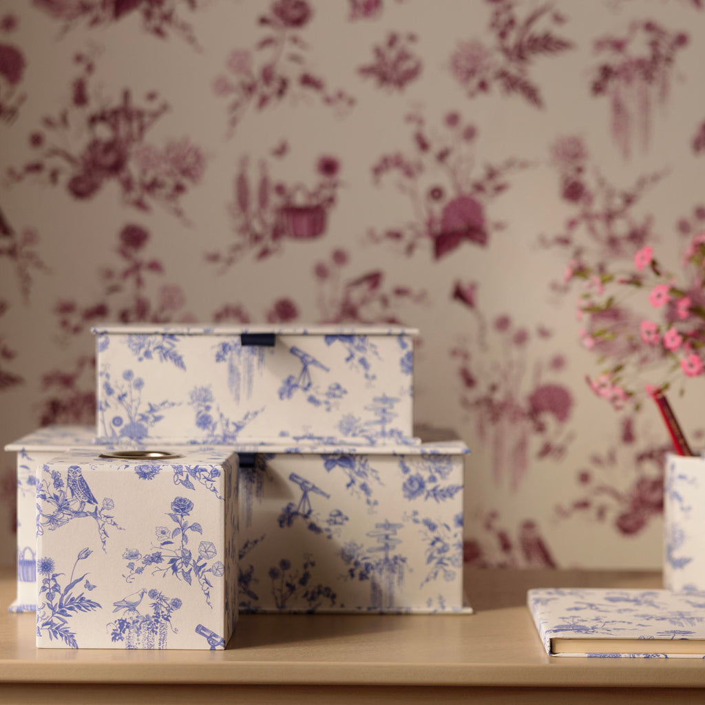 Three stacked fabric storage keepsake boxes with a blue and white toile de Jouy pattern, displayed on a light beige wooden table against a matching floral wallpaper background. 