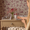 A beige wooden desk in a room with floral wallpaper, writing in a notebook. The desk has a vase of pink flowers and stationery, and the background features a matching floral pattern. Featuring a Delft Toile De Jouy pencil holder.