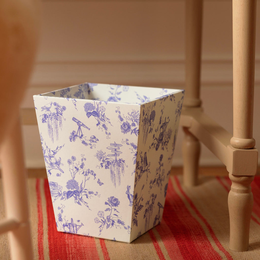 A wastepaper bin with a blue floral toile de Jouy pattern on a white background, styled on a red and beige striped rug next to a wooden table leg.