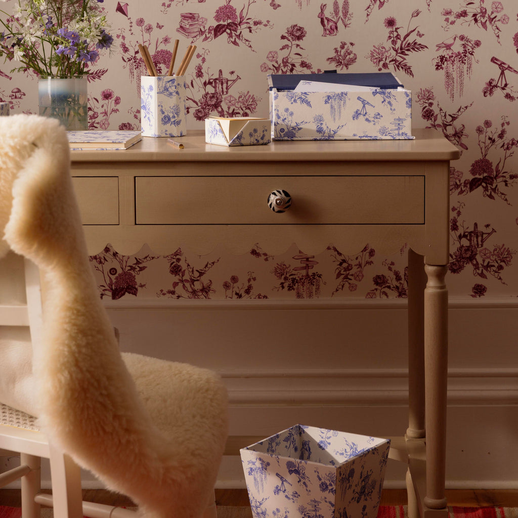 A styled interior scene featuring a wall covered in pink floral toile wallpaper, with a beige writing desk, a cream sheepskin chair, and matching blue-and-white patterned paper goods. Including a delft toile de jouy wastepaper bin and small A5 keep sake box.