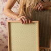 A woman holds two rectangular wooden picture frames against a floral wallpaper background. The frame in front has a pale sage green patterned insert, while the frame behind it has a dark brown patterned insert.