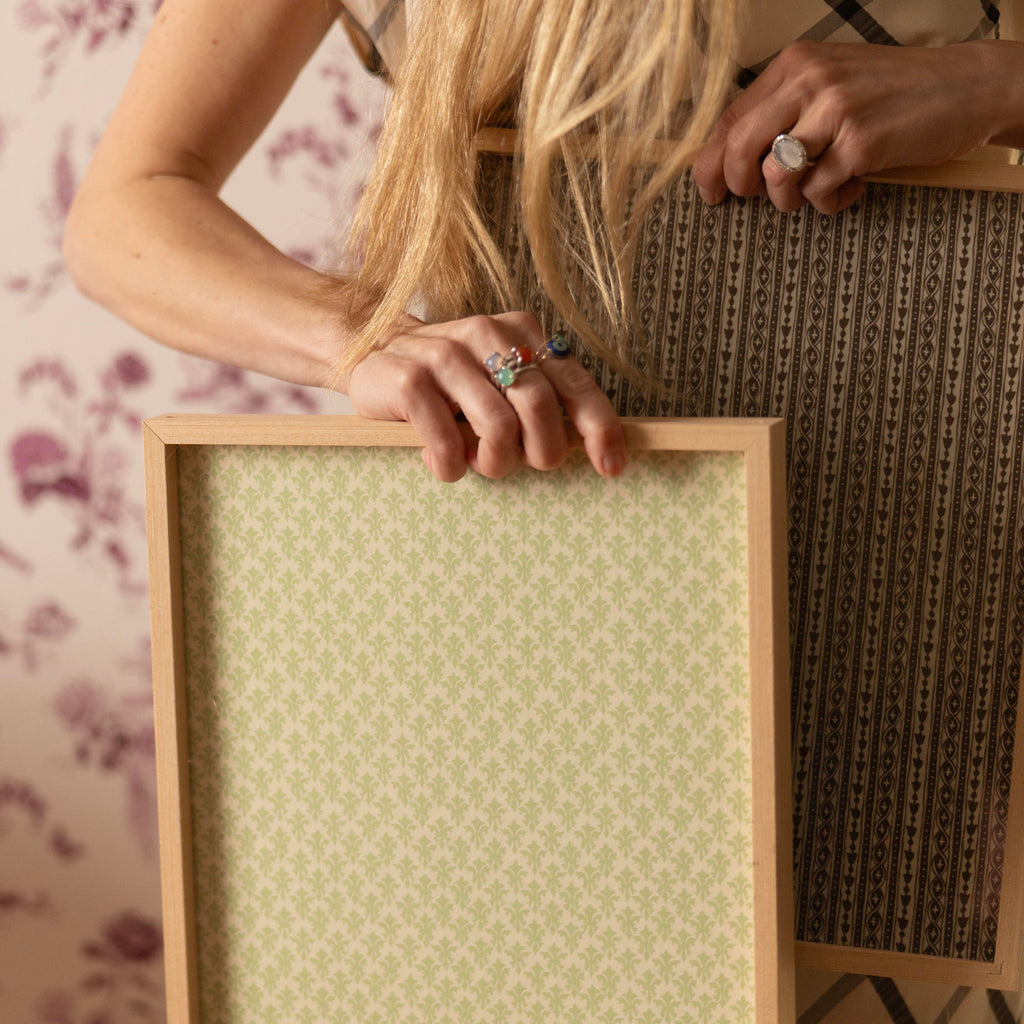A woman holds two rectangular wooden picture frames against a floral wallpaper background. The frame in front has a pale sage green patterned insert, while the frame behind it has a dark brown patterned insert.