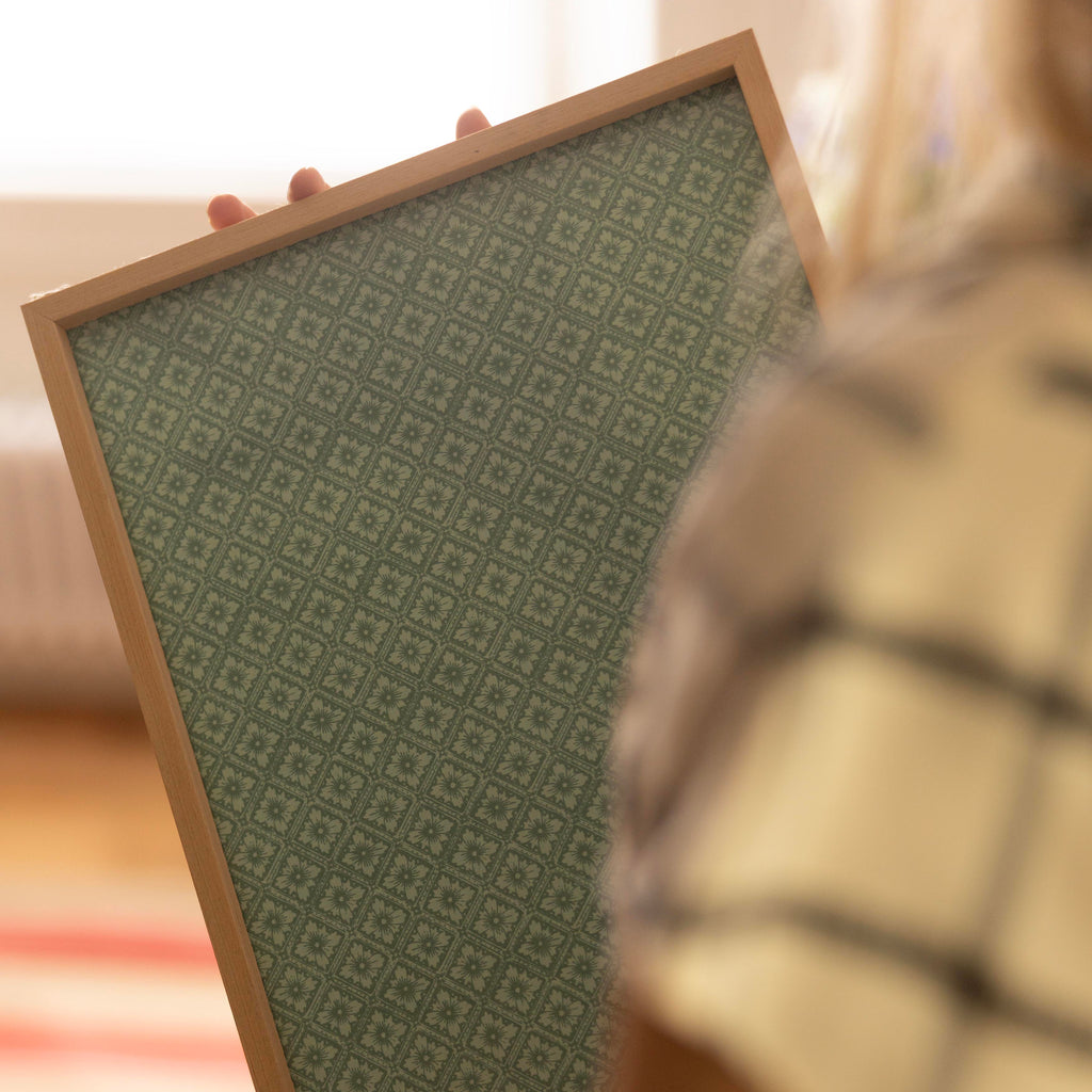 A person holds a rectangular mirror with a light wood frame and a green geometric patterned surface in a brightly lit room. green coloured rosace wallpaper house of petra