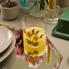 Glass with hand painted buttercup design filled with orange juice on a table with plates, mirrored placemats and a vase.