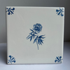 A square white ceramic tile with a blue floral design in the center and blue decorative corner motifs, photographed against a neutral gray background.