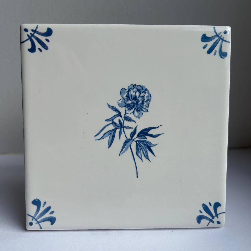 A square white ceramic tile with a blue floral design in the center and blue decorative corner motifs, photographed against a neutral gray background.