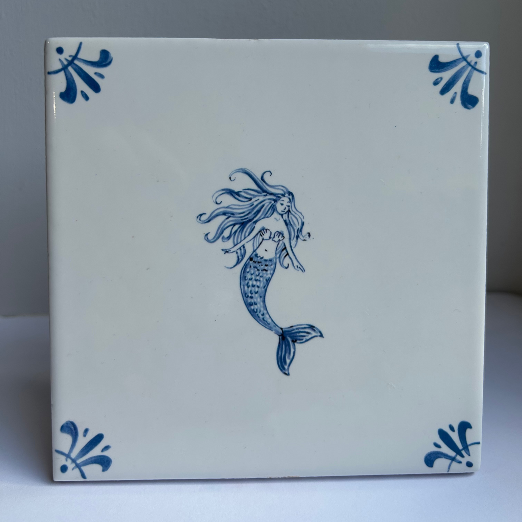 A square white ceramic tile featuring a blue hand-painted mermaid in the center, with blue decorative corner motifs, set against a neutral gray background.