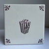 A square white ceramic tile featuring a manganese  print of a popcorn bucket with the words 'popcorn' and decorative flourishes in the corners, set against a neutral gray background.