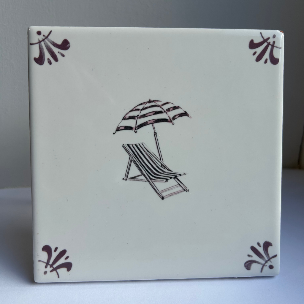 A square ceramic tile with a white glaze, featuring a central illustration of a striped beach umbrella and lounge chair, with matching decorative corner flourishes. Colour manganese 
