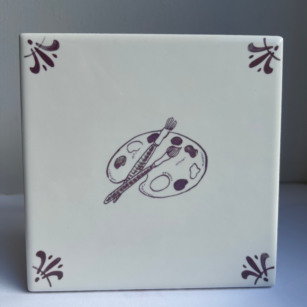 A square ceramic tile with a glossy off-white surface, featuring a central illustration of a paint palette and brush in manganese, along with matching decorative corner flourishes.