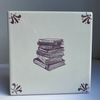 A square ceramic tile featuring a manganese illustration of a stack of books in the center, with manganese decorative flourishes in the four corners, set against a neutral gray background.