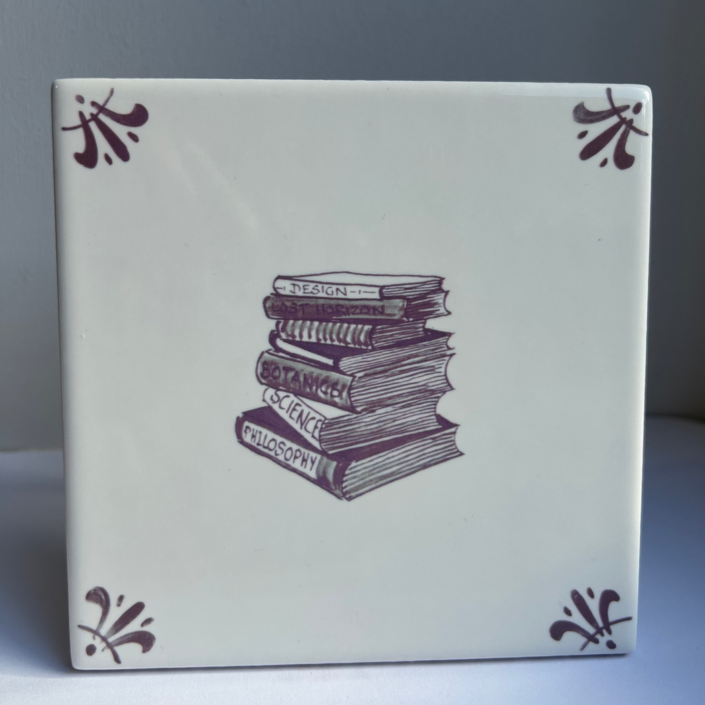 A square ceramic tile featuring a manganese illustration of a stack of books in the center, with manganese decorative flourishes in the four corners, set against a neutral gray background.