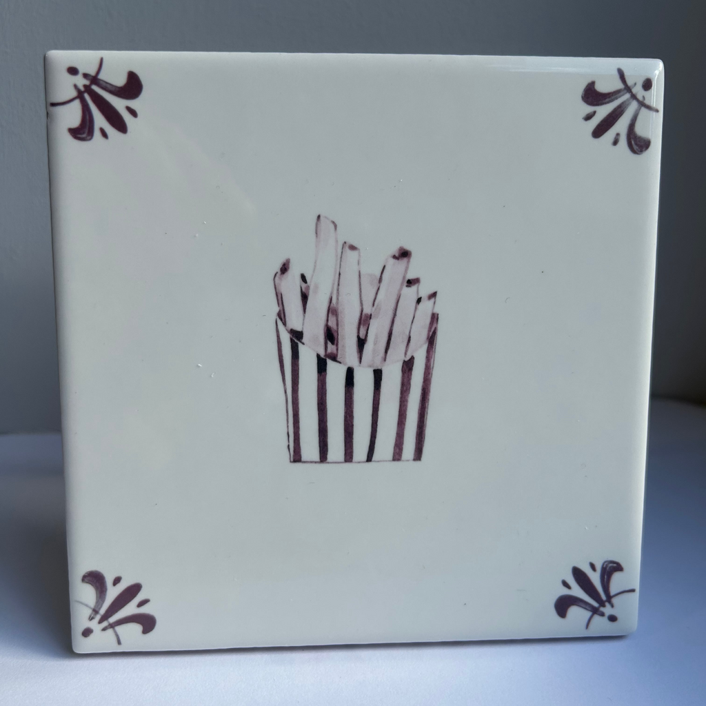 A square ceramic tile with a glossy white glaze, featuring a hand-painted illustration of french fries in a striped bucket in the center and matching manganeses corner flourishes.