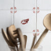 A decorative kitchen scene with white square tiles featuring a small red lobster motif in the center and matching red floral corner accents. Blurred wooden utensils and a wooden spoon are visible in the foreground.